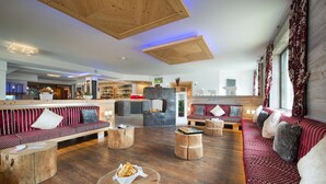 A spacious lounge area with wooden flooring, multiple seating options including sofas and round tables, and a visible bar area in the background. - Hotel Cristallo (Moena)