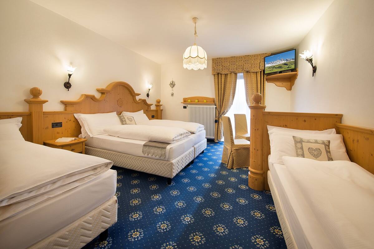 A hotel room with a large bed, two smaller beds, a wooden headboard, a nightstand, a lamp, a television, and a patterned carpet.