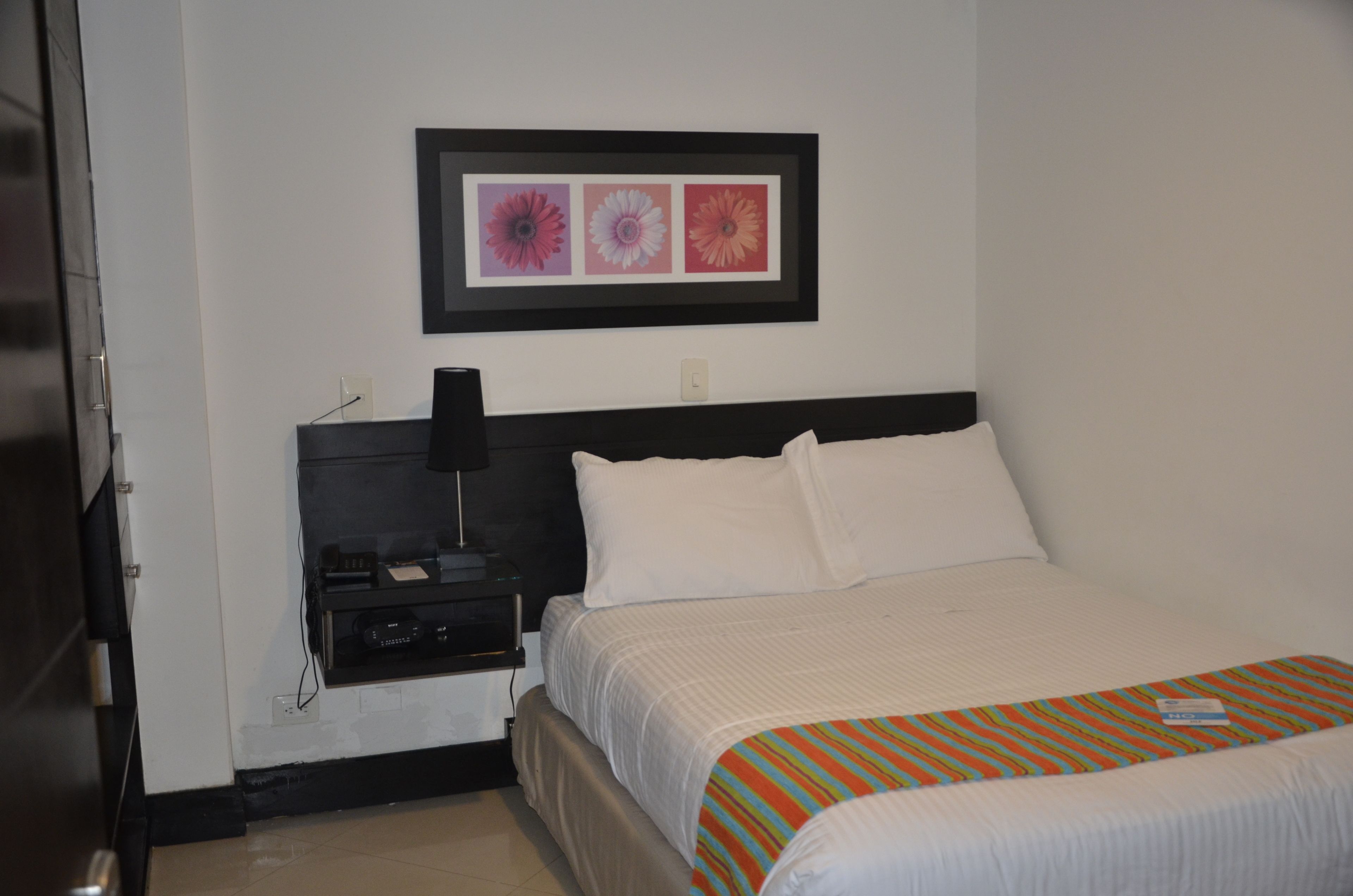 A hotel room with a bed, a nightstand, a framed picture on the wall, and a door.