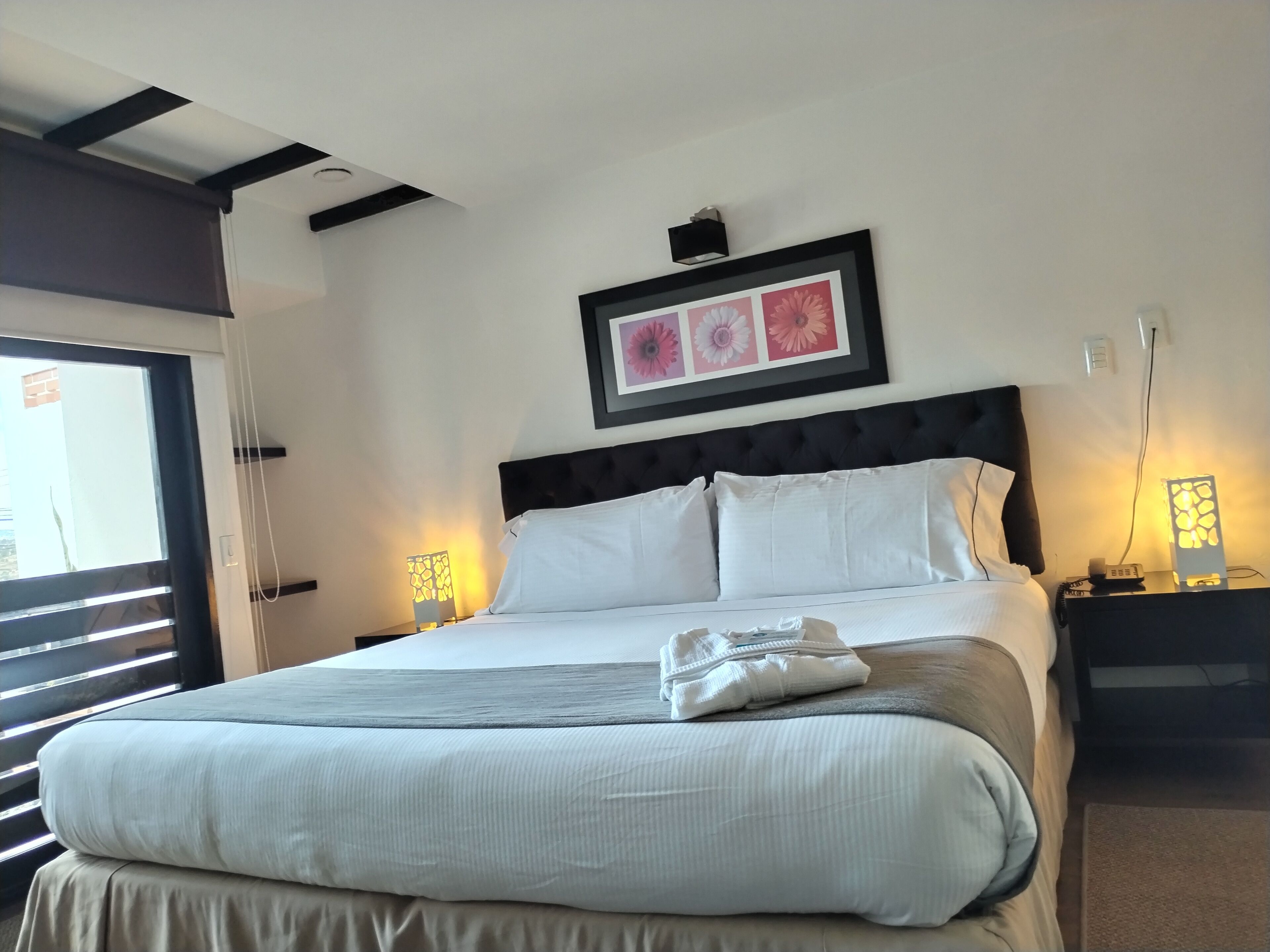 A neatly made bed with a grey and white duvet, a black headboard, and a framed floral artwork above it.