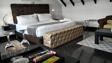 A modern hotel room with a large bed, a tufted bench, a bedside table with glasses and snacks, and a framed flower artwork on the wall.