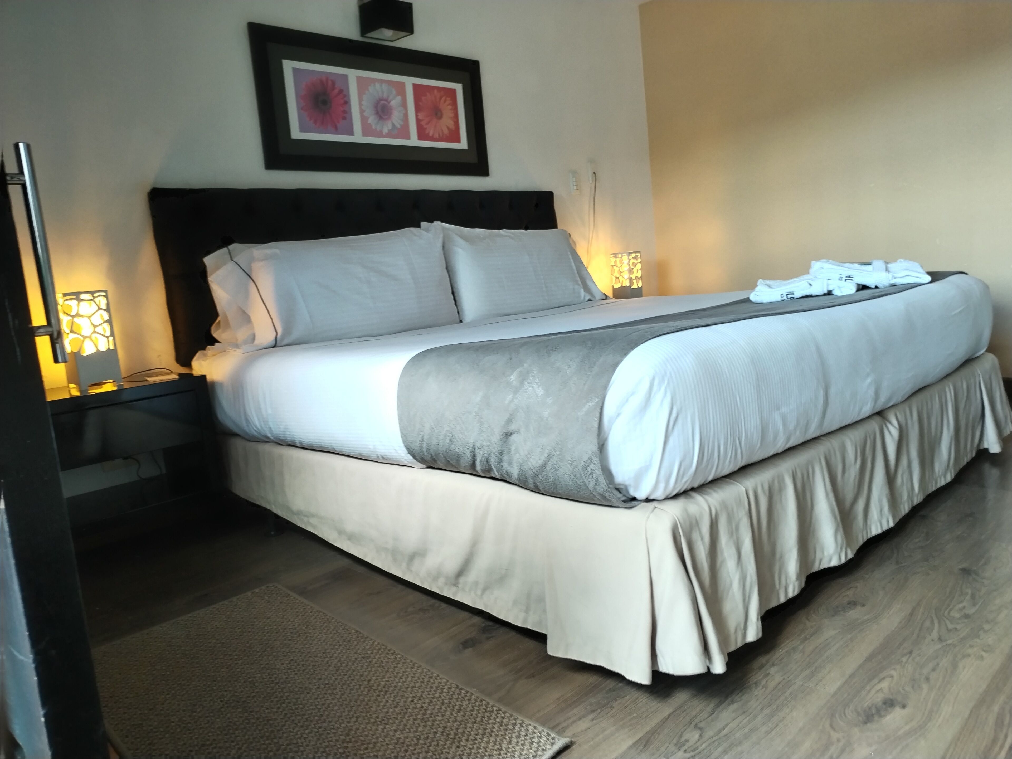 A neatly made bed with a headboard, a bedside table with a lamp, and a framed picture on the wall.