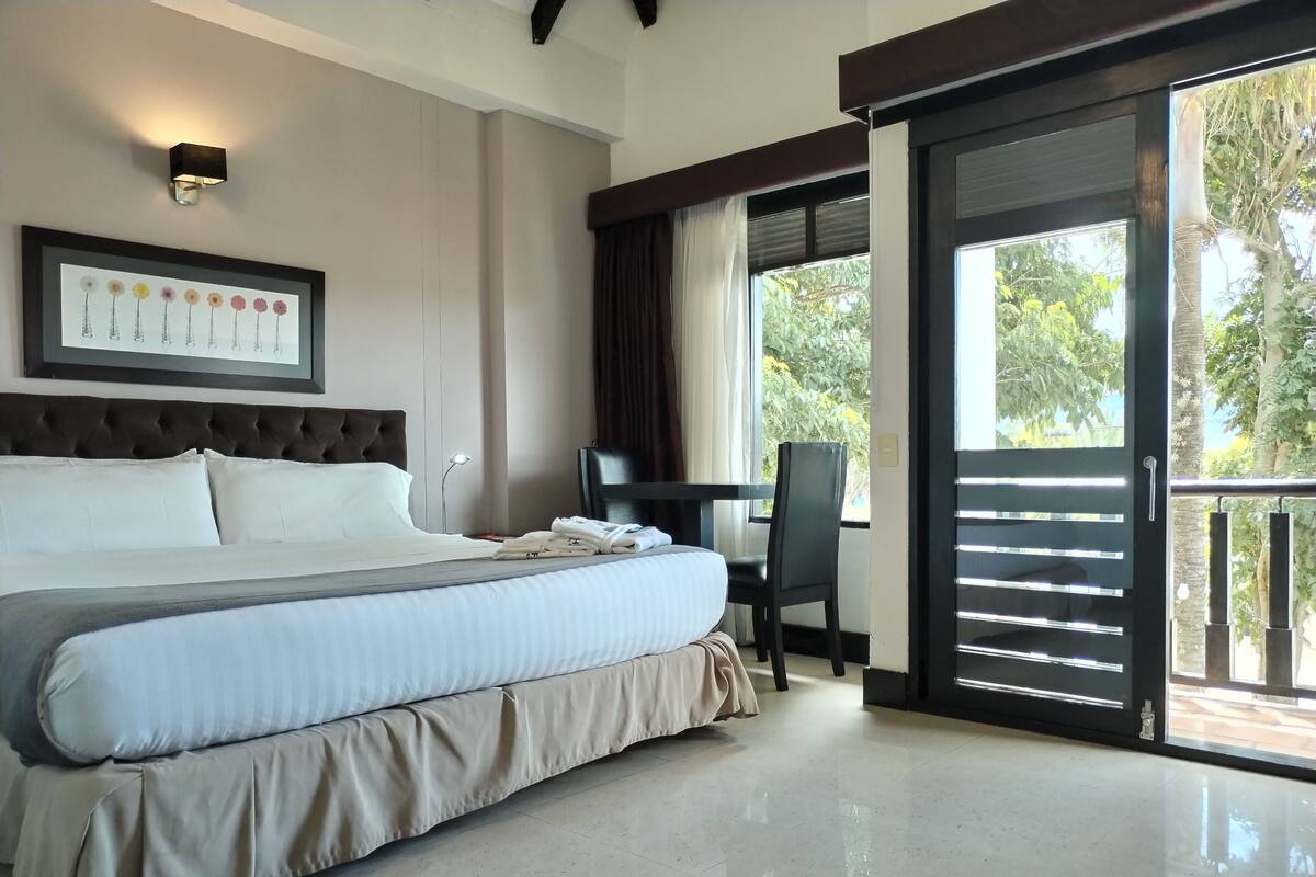 A hotel room with a large bed, a desk, and a balcony with a view of greenery.