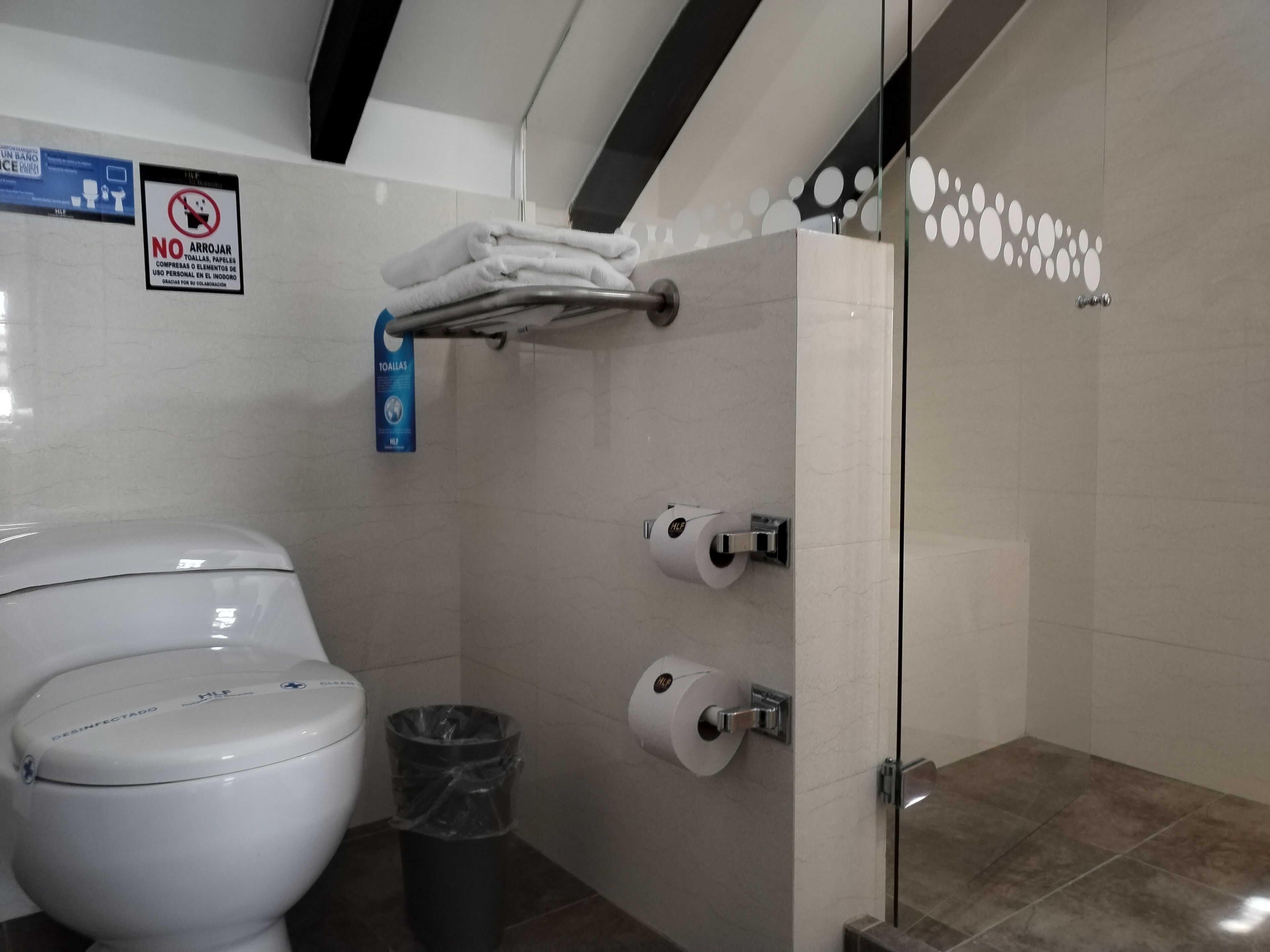 A modern bathroom with a toilet, two paper towel dispensers, a trash bin, and a glass shower enclosure.