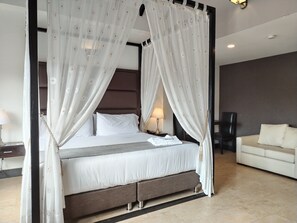 Una cama con dosel con ropa blanca y un dosel, una mesita de noche con lámpara y un sofá blanco en una habitación de hotel moderna.