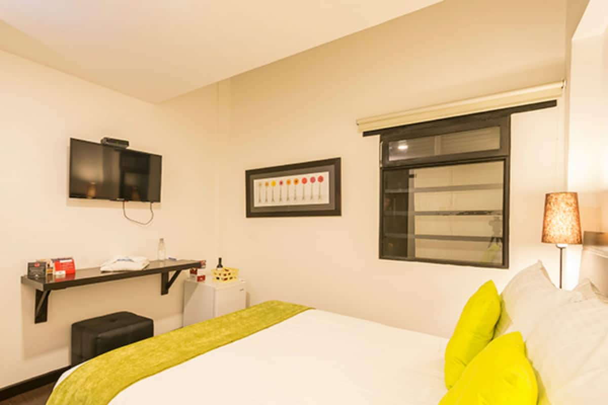 A hotel room with a bed, a television, a small desk, a window, and a lamp.