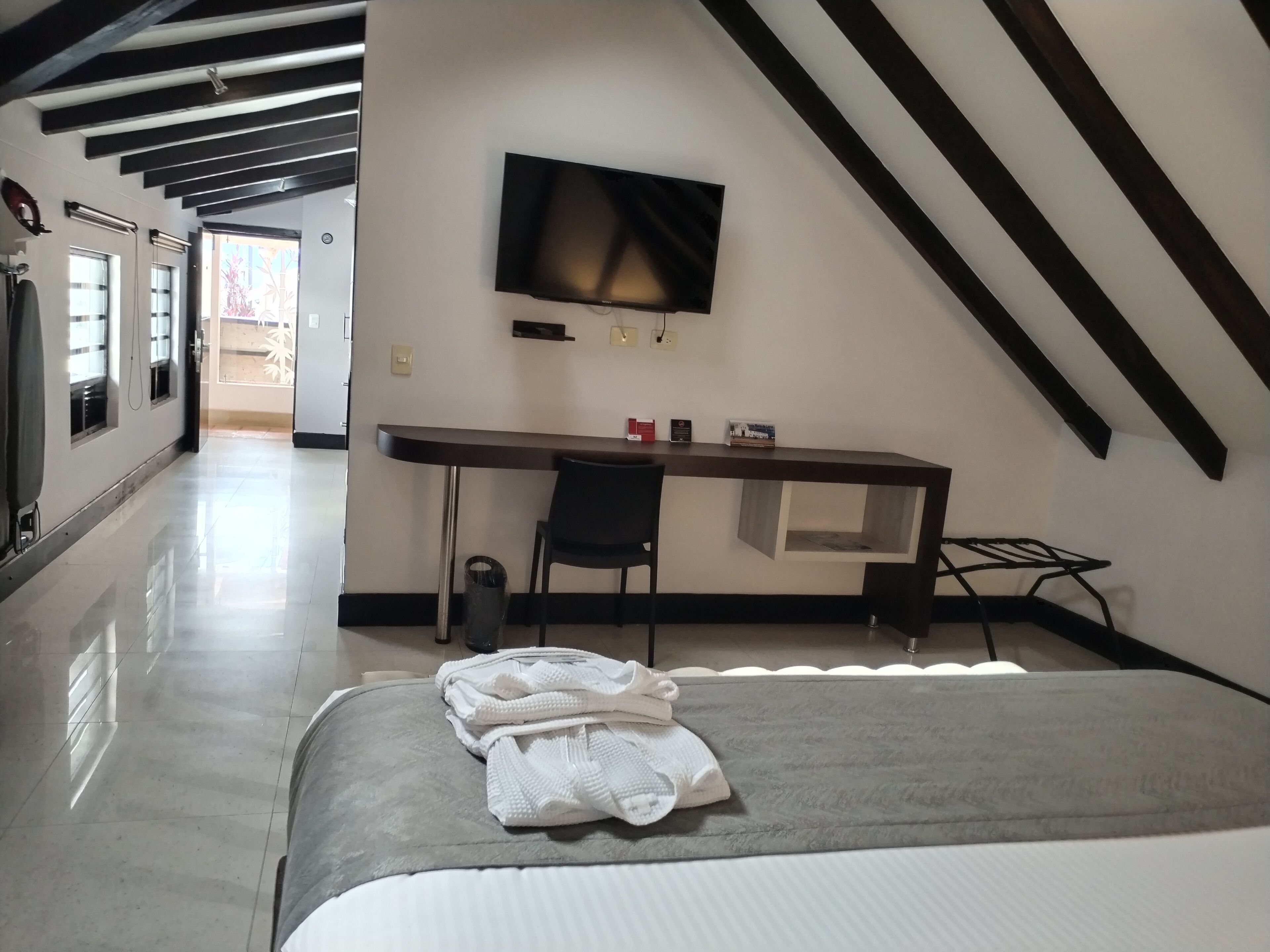 Una habitación de hotel con cama, escritorio, silla, televisor montado en la pared y ventana.