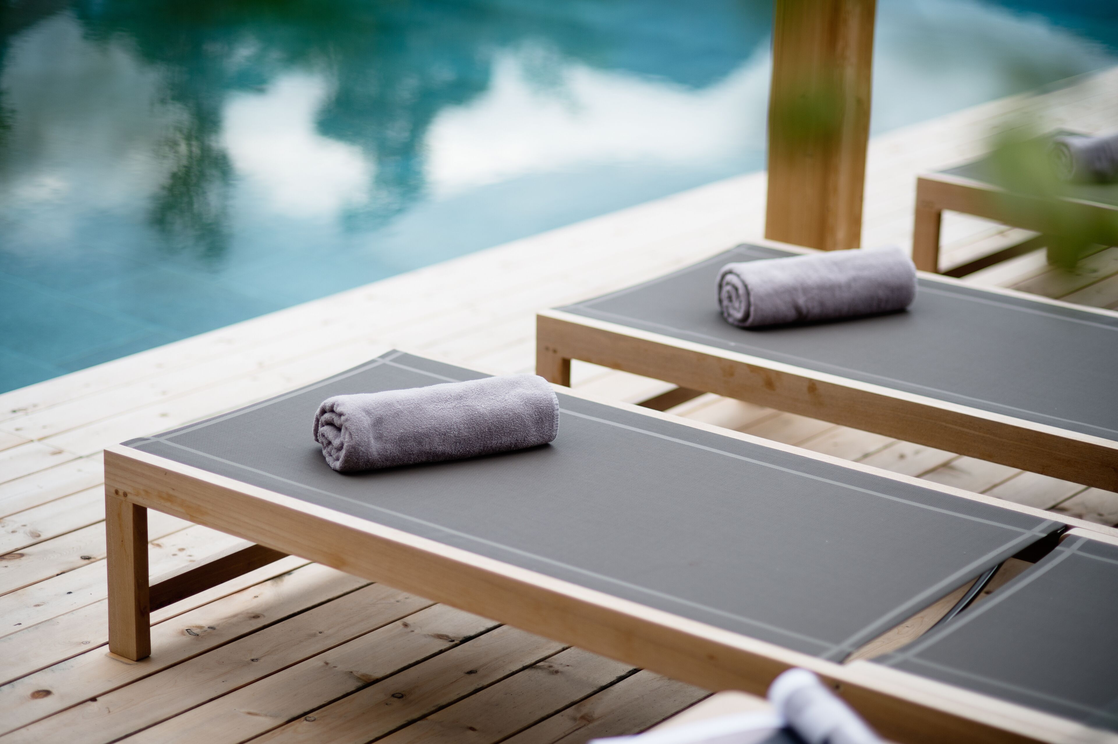 Zwei Holzliegestühle mit grauen Kissen und Handtüchern auf einer Holzterrasse an einem Pool.