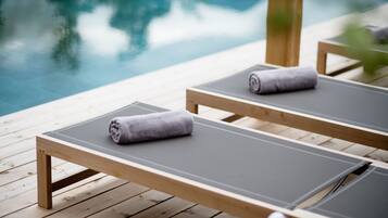 Zwei Holzliegestühle mit grauen Kissen und Handtüchern auf einer Holzterrasse an einem Pool.