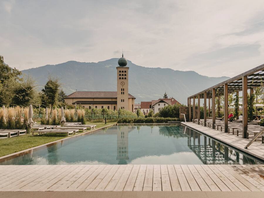 Un área de piscina con terraza de madera, sillas de descanso y un edificio con una torre del reloj al fondo.