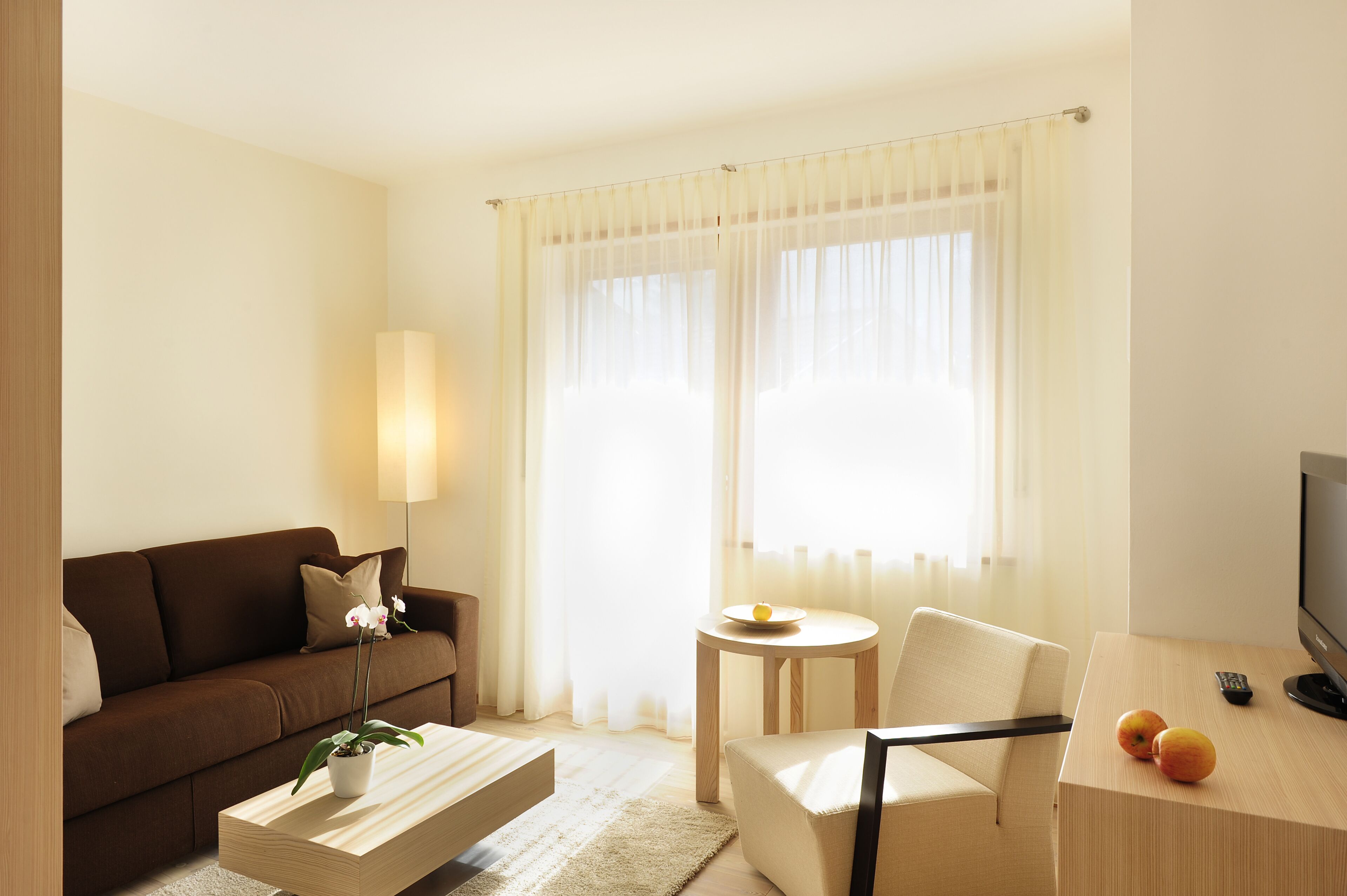 A modern hotel room with a brown sofa, a small round table with a lamp, a TV, and a wooden coffee table with a potted plant and fruit.