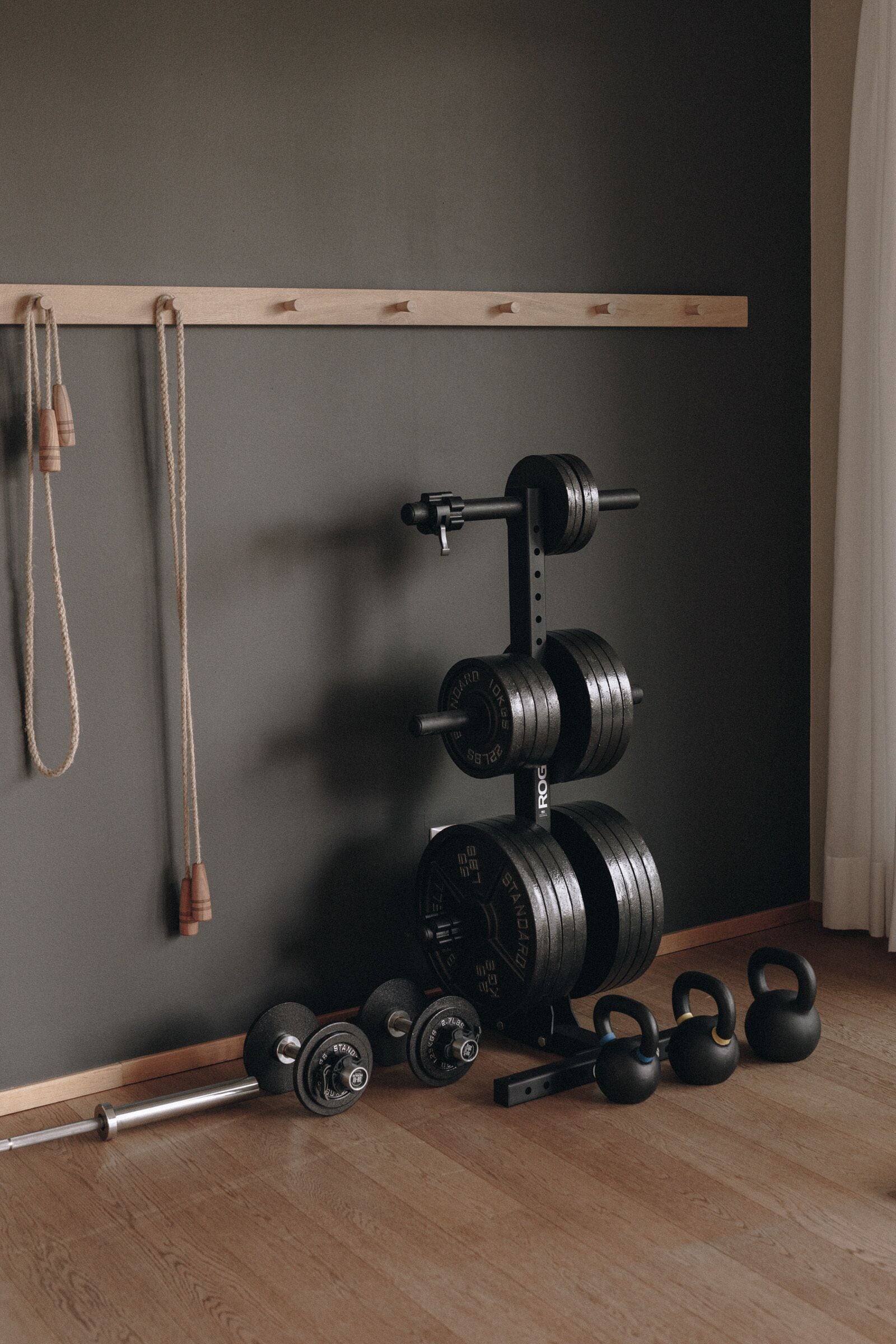 A home gym with a barbell rack, kettlebells, dumbbells, and resistance bands against a dark wall.