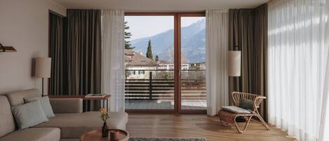 A modern living room with a sofa, coffee table, and a wicker chair. A large window offers a view of mountains and a balcony.