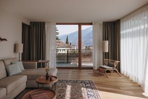 Una sala de estar moderna con un sofá, una mesa de centro y una silla de mimbre. Un ventanal ofrece vistas a montañas y a un balcón.