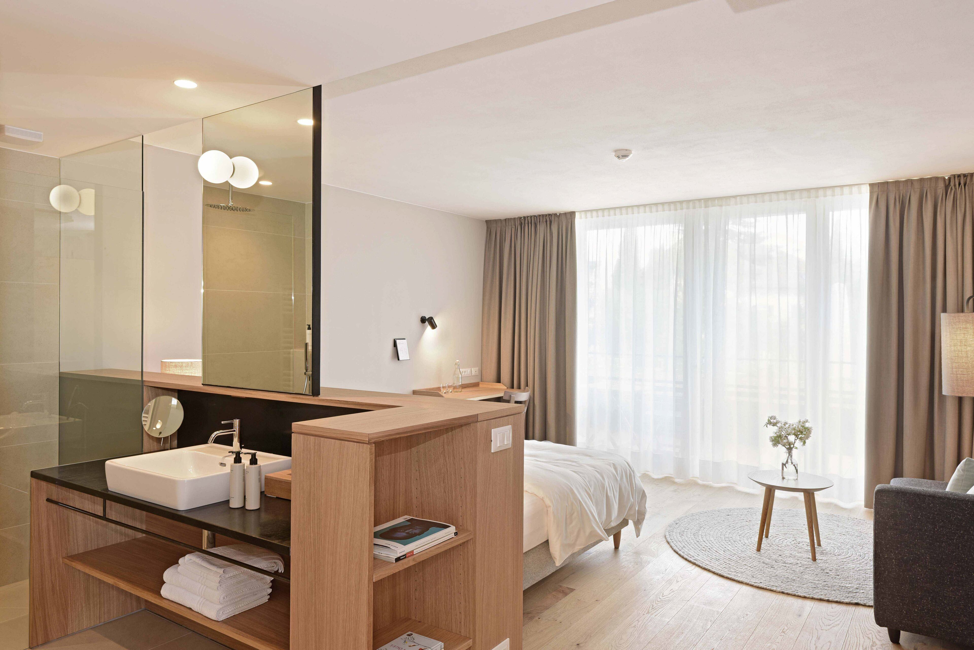 Une chambre d’hôtel moderne avec un grand miroir, une coiffeuse en bois, un lit, un tapis rond et une petite table sur laquelle est posé un vase.