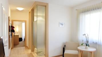 Una habitación de hotel moderna con suelo de madera, un separador de vidrio, un sofá beige, una pequeña mesa redonda con una planta y un ventanal con cortinas de gasa.