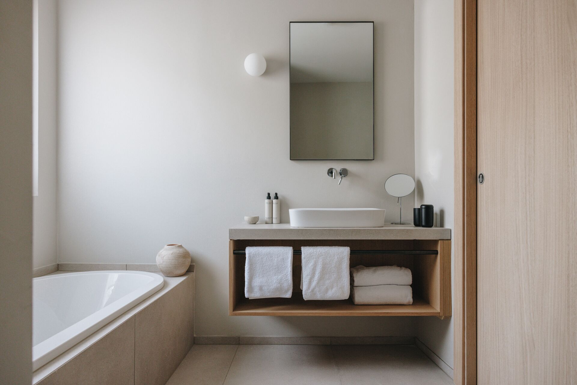 A modern bathroom with a freestanding bathtub, a white sink, a mirror, and a wooden shelf with towels.