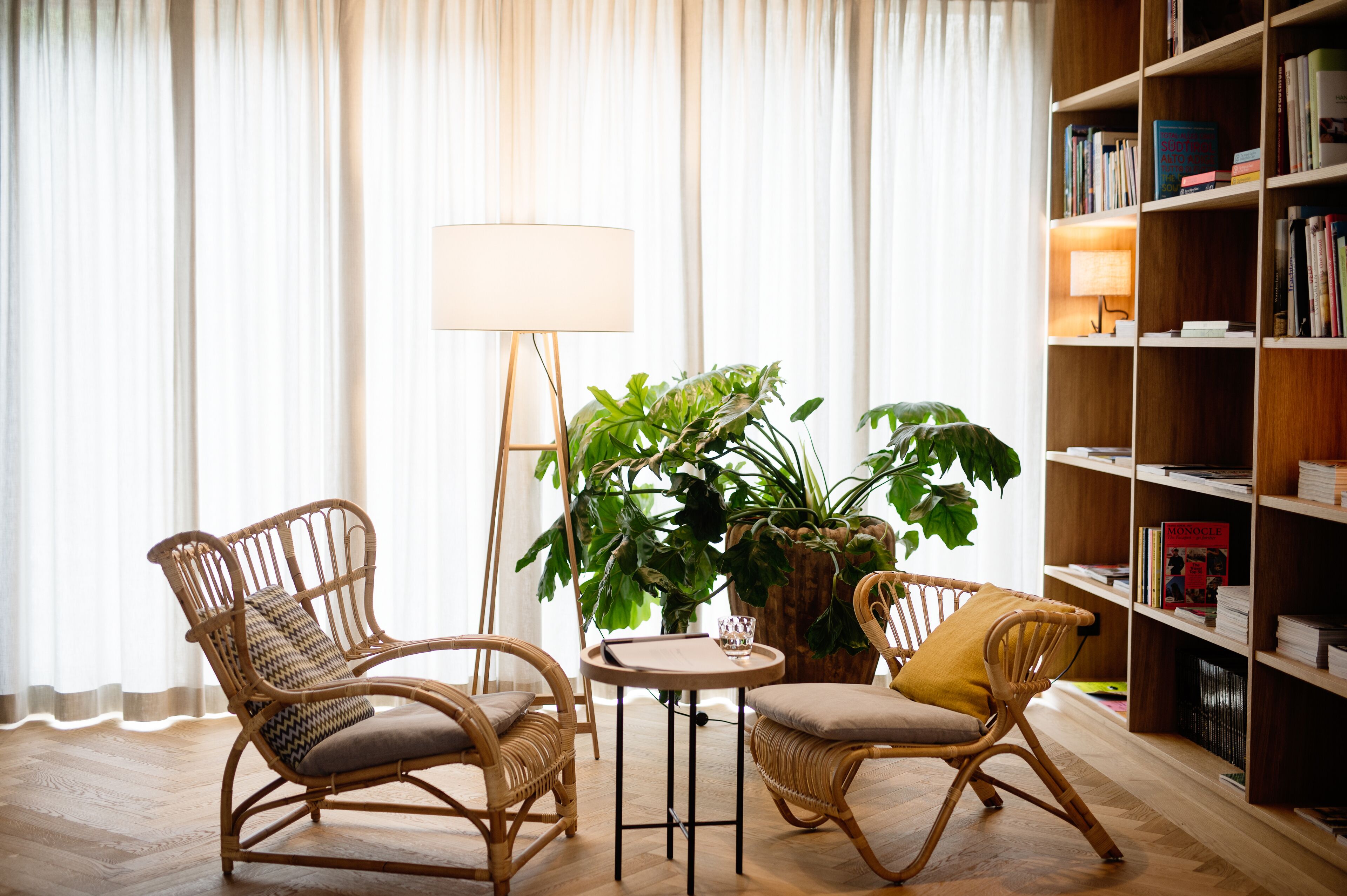 Una habitación con dos sillones de mimbre, una pequeña mesa redonda, una lámpara de pie, una planta grande en maceta y una estantería llena de libros.