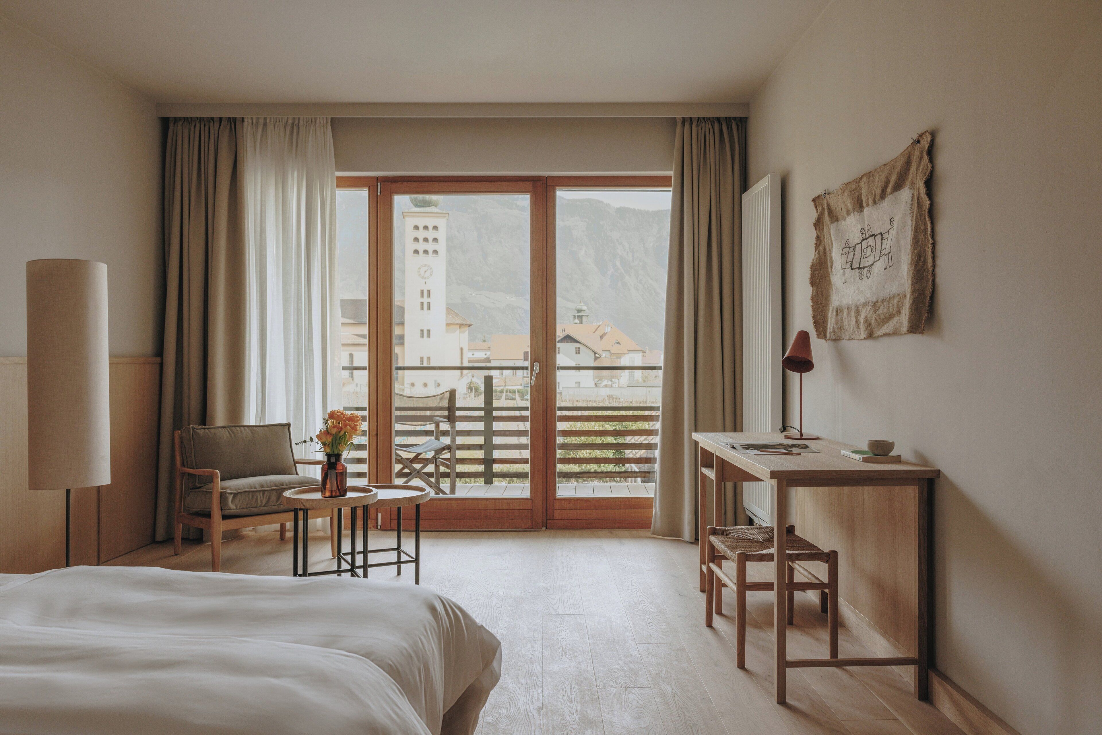 Une chambre d’hôtel avec un lit, un bureau, une chaise, une lampe et une vue sur une montagne et des bâtiments, visible par la fenêtre.