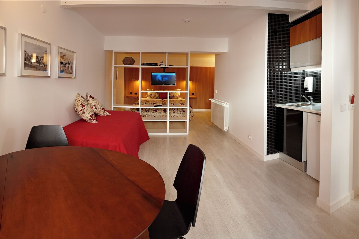 Ein modernes Hotelzimmer mit einem Holztisch, einem Bett mit roter Bettwäsche, einem Fernseher im Hintergrund und einer Küchenzeile.