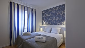 Une chambre d’hôtel avec deux lits, une grande fenêtre ornée de rideaux bleus et blancs, et un papier peint à motifs floraux sur le mur.