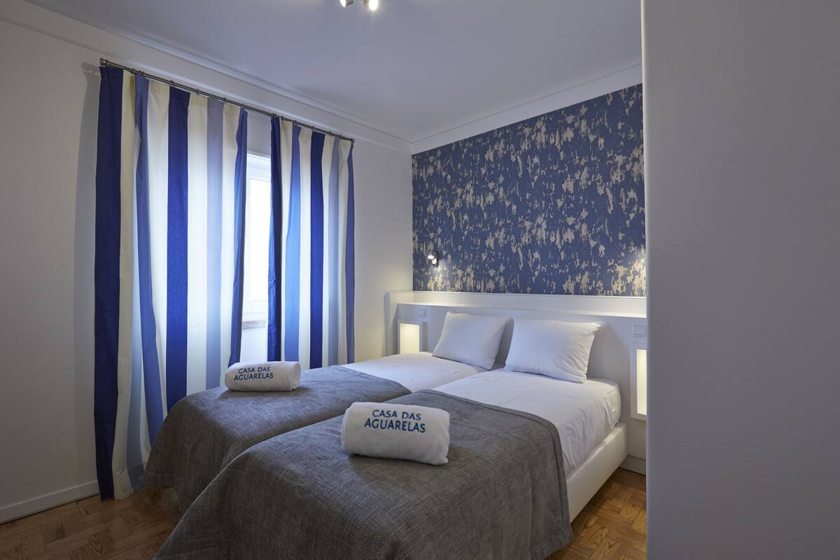 A hotel room with two beds, a large window with blue and white curtains, and a floral wallpaper on the wall.
