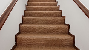 Un escalier recouvert de moquette, doté de rampes en bois, mène à une pièce lumineuse où l’on aperçoit une chaise au loin.