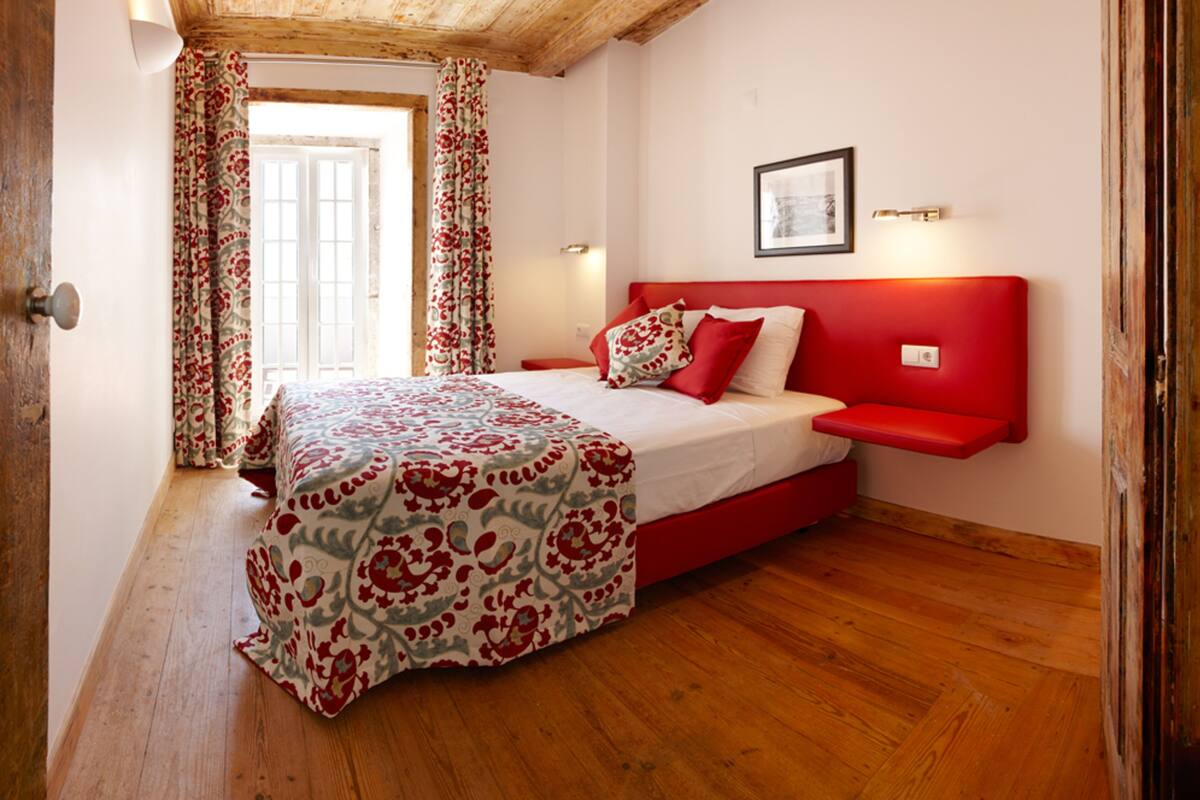 A bedroom with a bed, a red headboard, a window with curtains, and a framed picture on the wall.