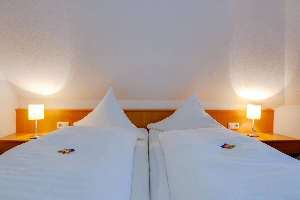 Hotellihuone, jossa on kaksi erillistä sänkyä, ja jokaisessa sängyssä on tyyny ja pieni esine. Sängyn vieressä on kaksi puista yöpöytää, ja niiden päällä on lamput.