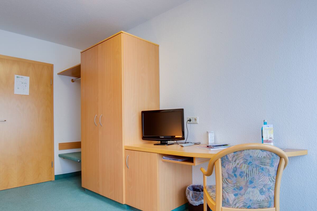 Huoneessa on puinen vaatekaappi, työpöytä, jossa on tietokone, ja kuviollinen tuoli.