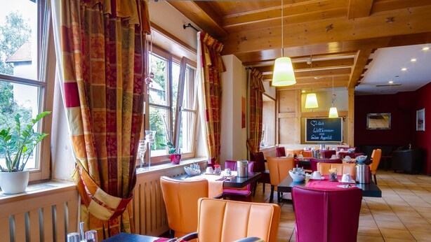 Un restaurant avec des poutres en bois, des chaises recouvertes d’un tissu orange et rouge, et un menu écrit à la craie sur un tableau noir.