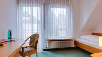 A small, well-lit room with a single bed, a wooden desk, a chair, and a radiator under a window.