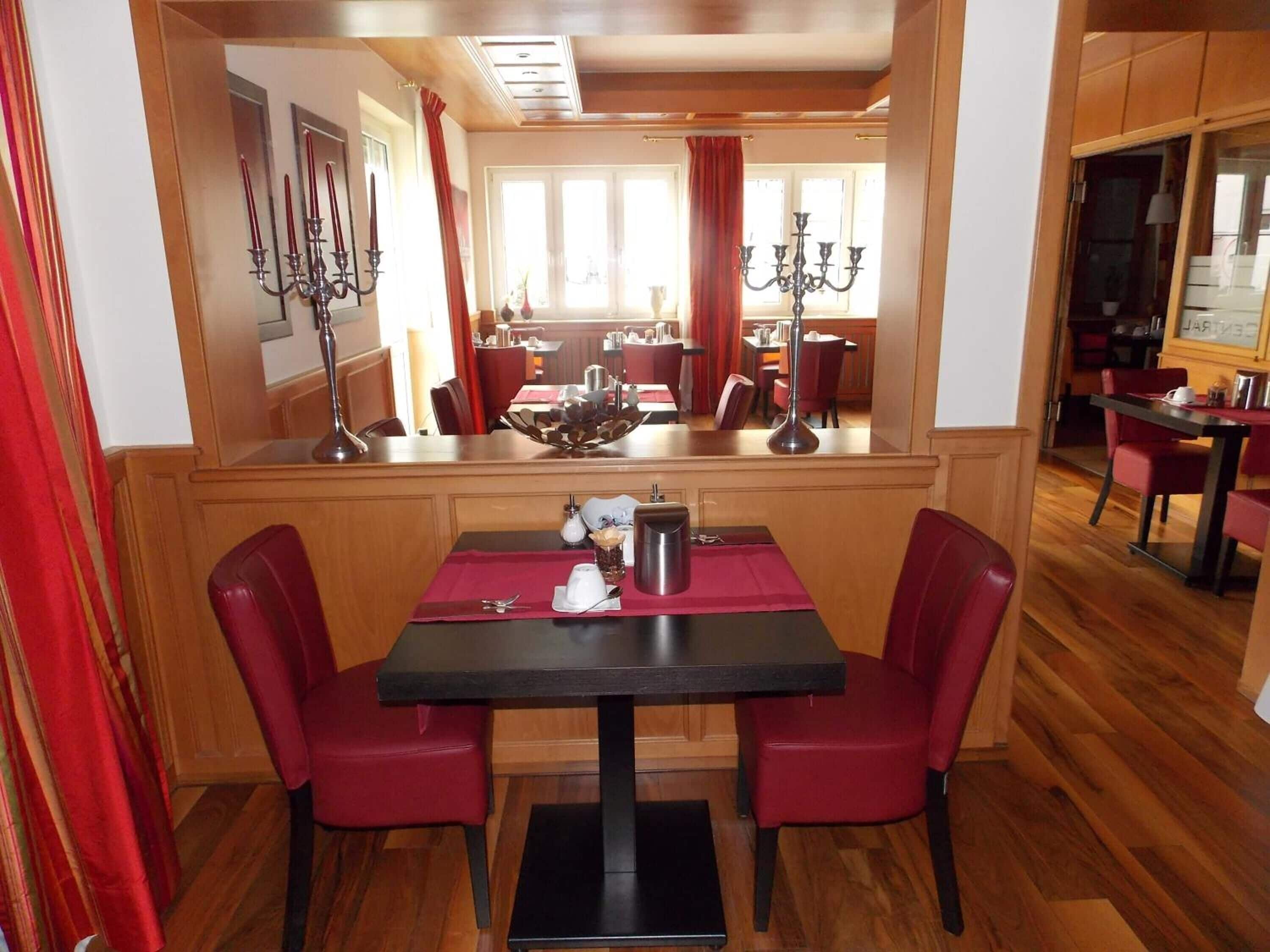 A dining area with a table and two chairs, a chandelier, and a view into a dining room with multiple tables and chairs.