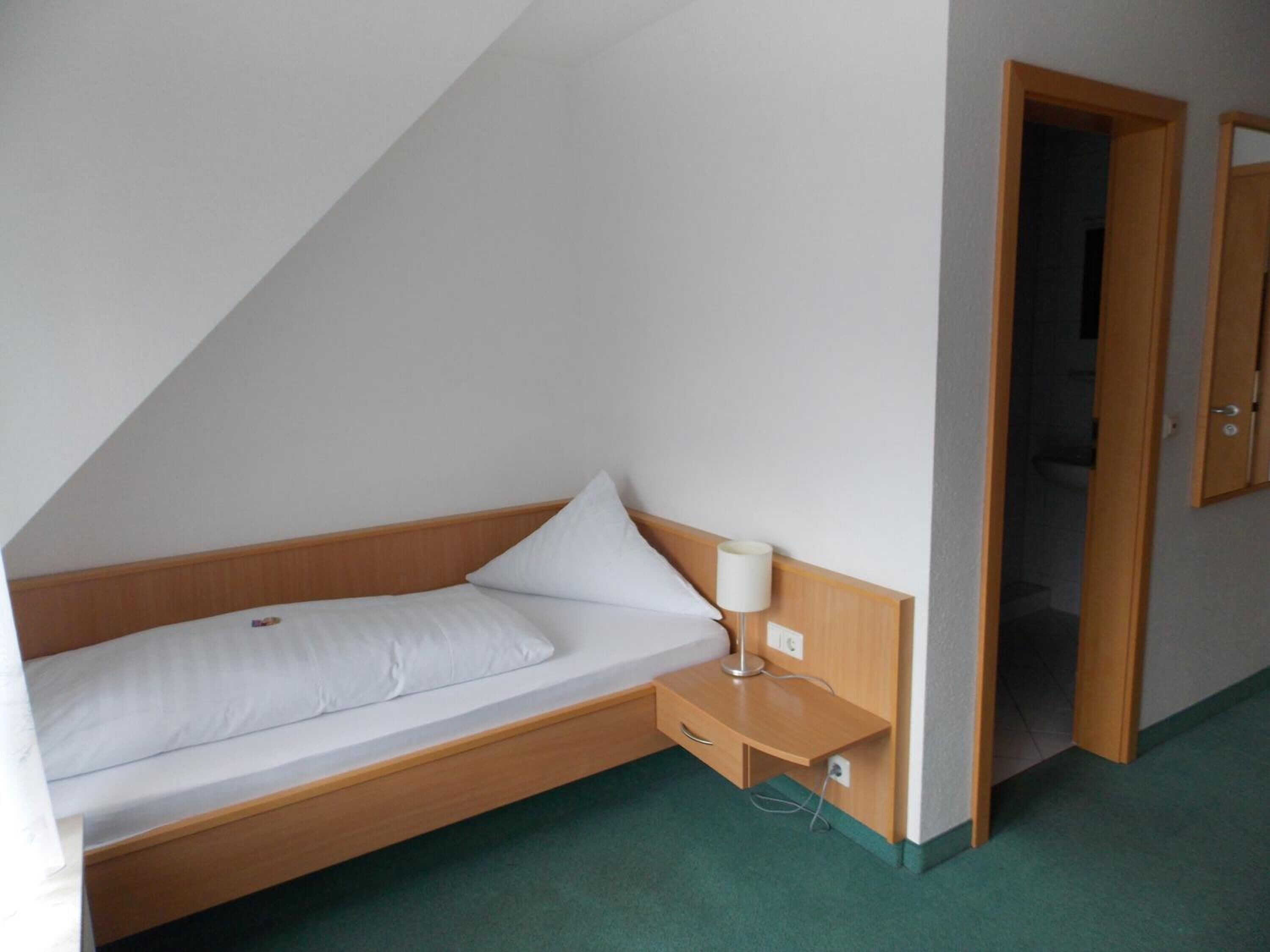 A small, single-bed room with a wooden headboard, a bedside table with a lamp, and a door leading to a bathroom.