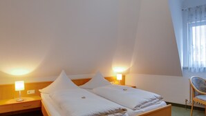 Une chambre d’hôtel avec un lit double, deux lampes de chevet, une table de chevet en bois, une chaise et une fenêtre avec des rideaux légers.