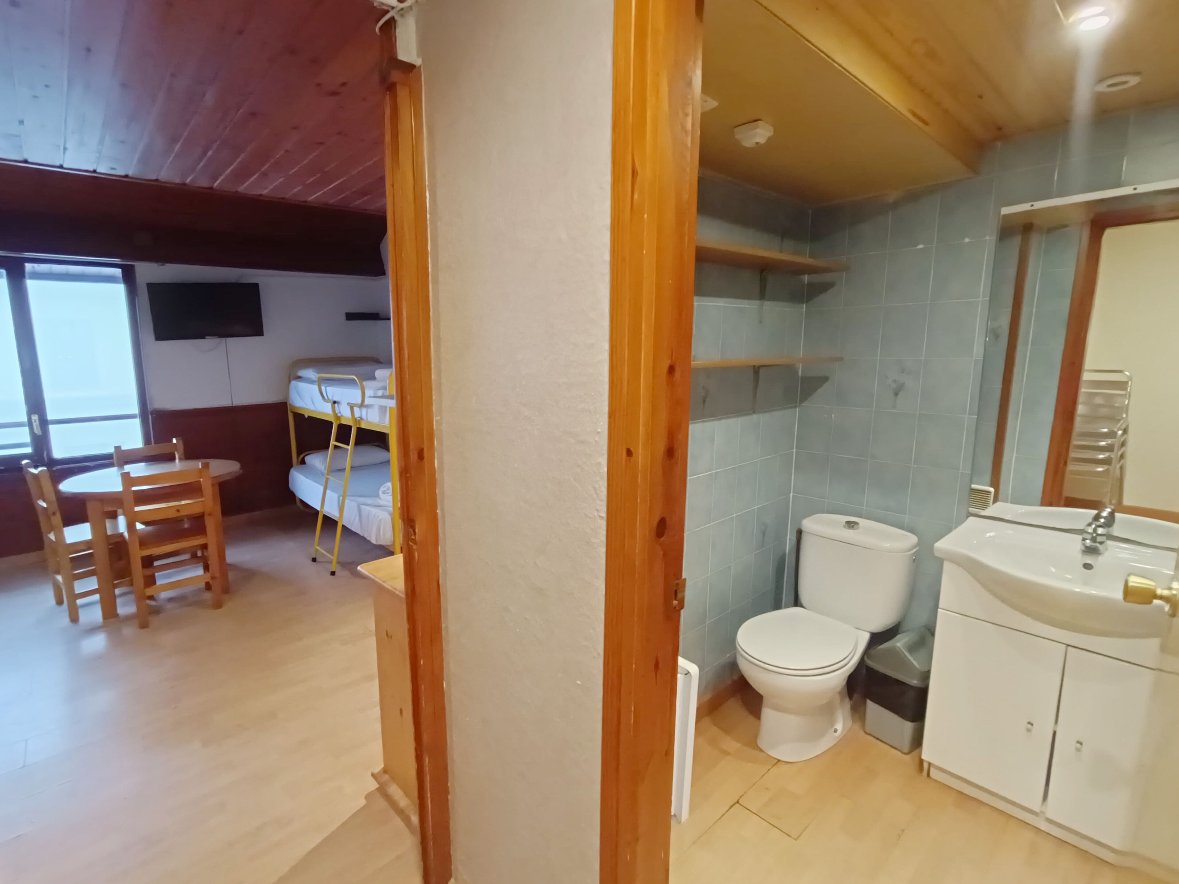 A room with a wooden ceiling, a bunk bed, a small table with chairs, and a bathroom with a toilet and sink.