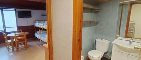 A room with a wooden ceiling, a bunk bed, a small table with chairs, and a bathroom with a toilet and sink.