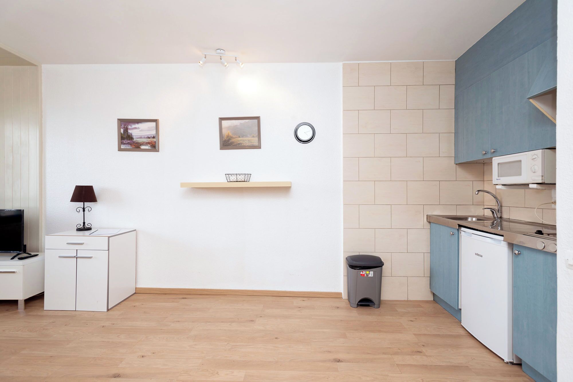 Cocina compacta con microondas, pileta y alacenas. Hay un cubo de basura pequeño y una repisa con una cesta.
