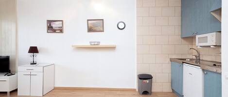 Cocina compacta con microondas, pileta y alacenas. Hay un cubo de basura pequeño y una repisa con una cesta.