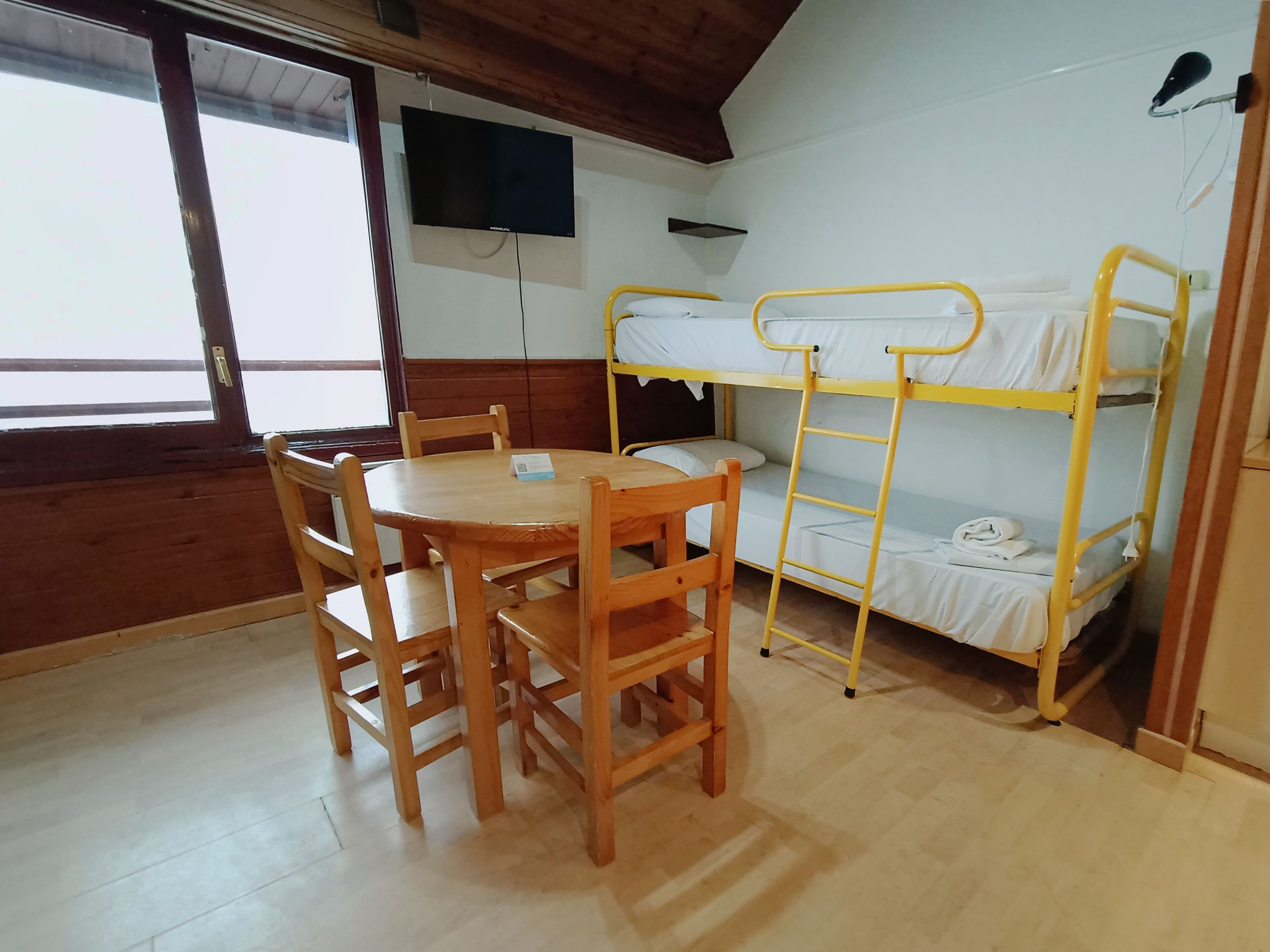 A room with bunk beds, a wooden table with chairs, a TV mounted on the wall, and a window providing natural light.