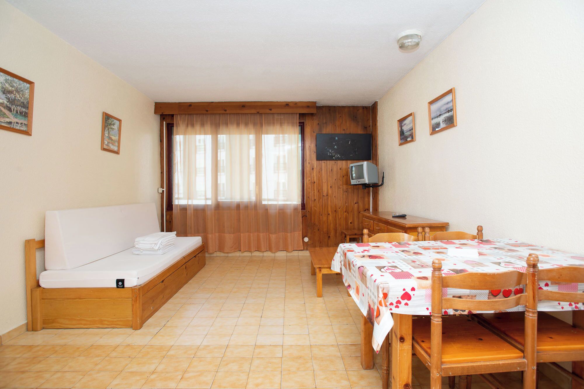 A small, cozy room with a bed, a desk, and a chair. There is a television on the wall and a window with curtains.