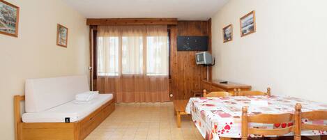 A small, cozy room with a bed, a desk, and a chair. There is a television on the wall and a window with curtains.