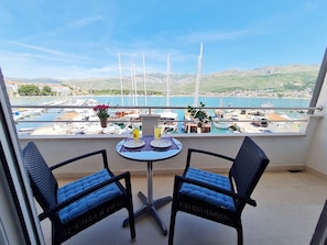 Un balcón con dos sillas y una mesa, con vista a un puerto deportivo con barcos y un paisaje montañoso.