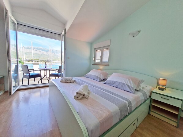 Quarto com cama, mesa de cabeceira, abajur e vista para uma varanda com cadeiras e mesa.