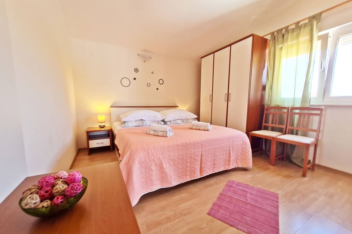 A neatly arranged bedroom with a bed, bedside tables, a wooden chair, a small table with a bowl, and a window with curtains.