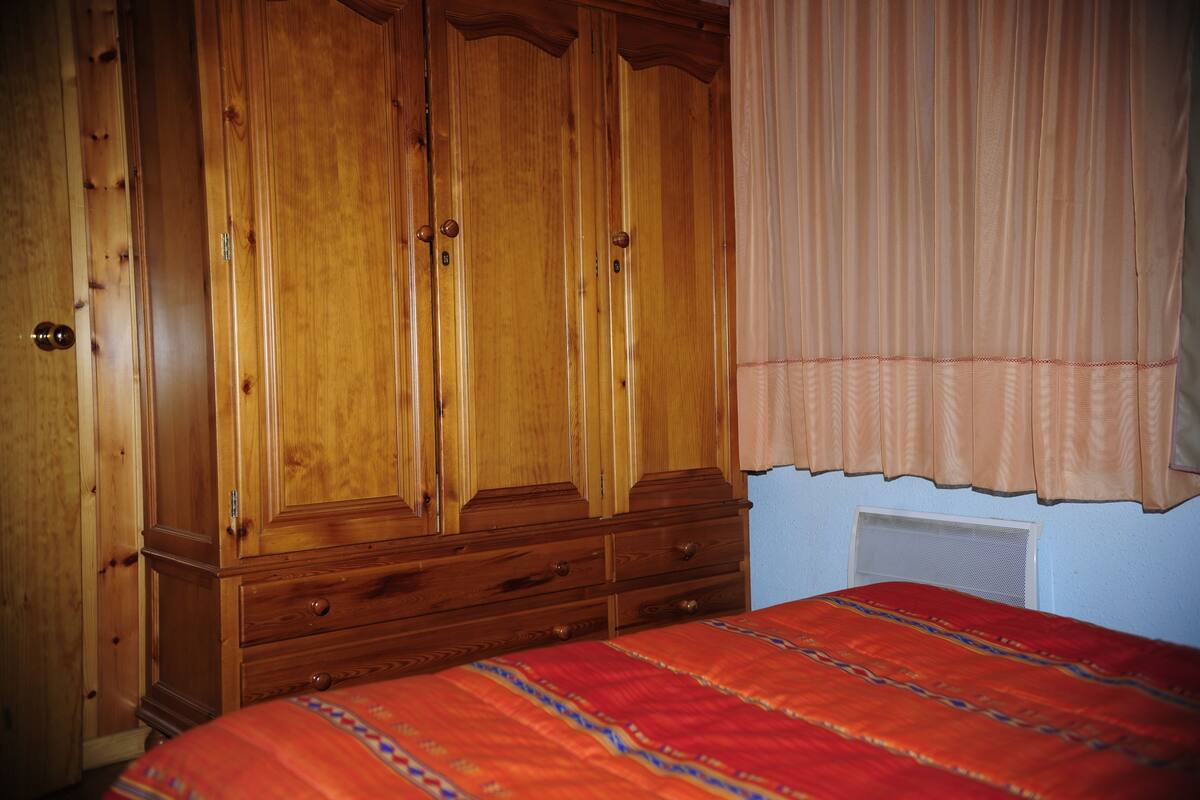 A bedroom with a wooden wardrobe, a bed with a patterned bedspread, and a window with a beige curtain.