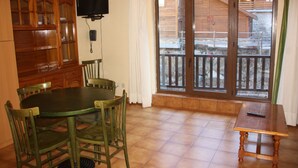 A dining area with a round table and chairs, a wooden cabinet, a television, and a sliding glass door leading to an outdoor area. - Deusol 3000 (Soldeu)