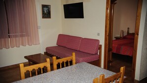 A small room with a wooden table and chairs, a red patterned sofa, a television mounted on the wall, and a door leading to another room with a bed. - Deusol 3000 (Soldeu)