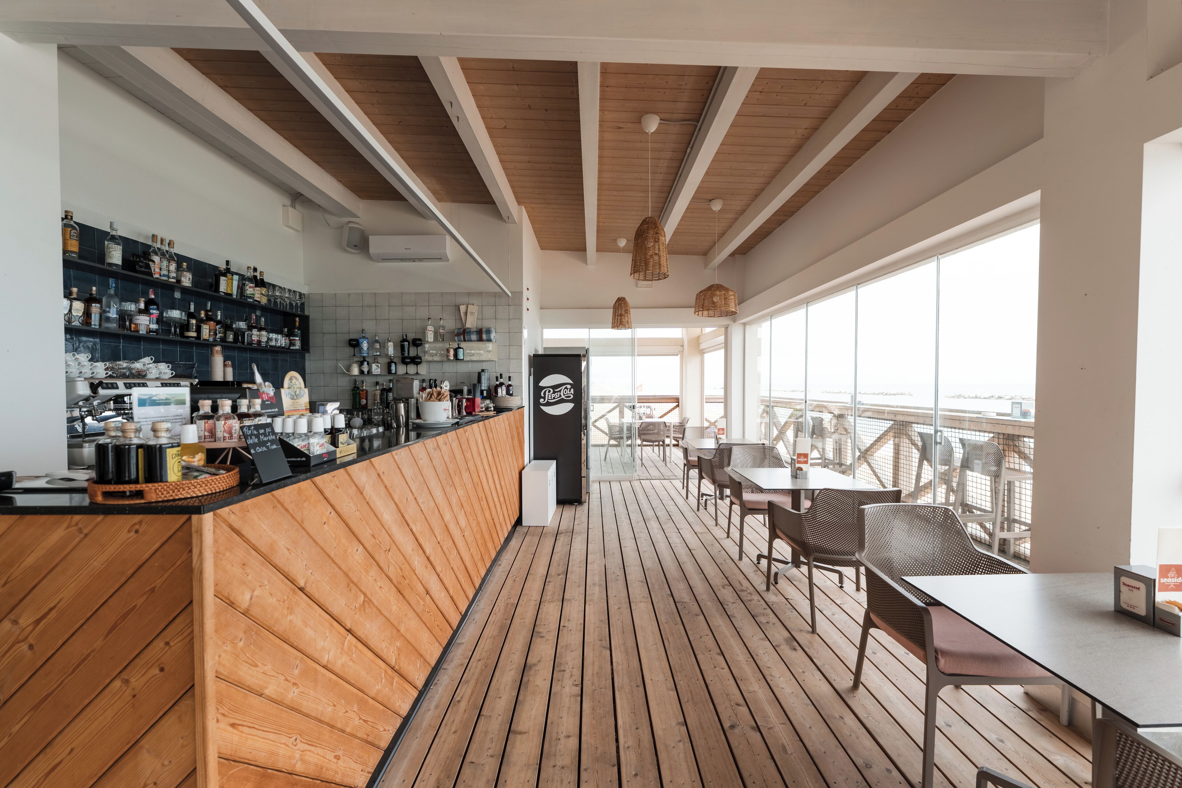 Eine Bar mit Holzboden, einer Theke voller Flaschen und Blick durch große Fenster nach draußen.
