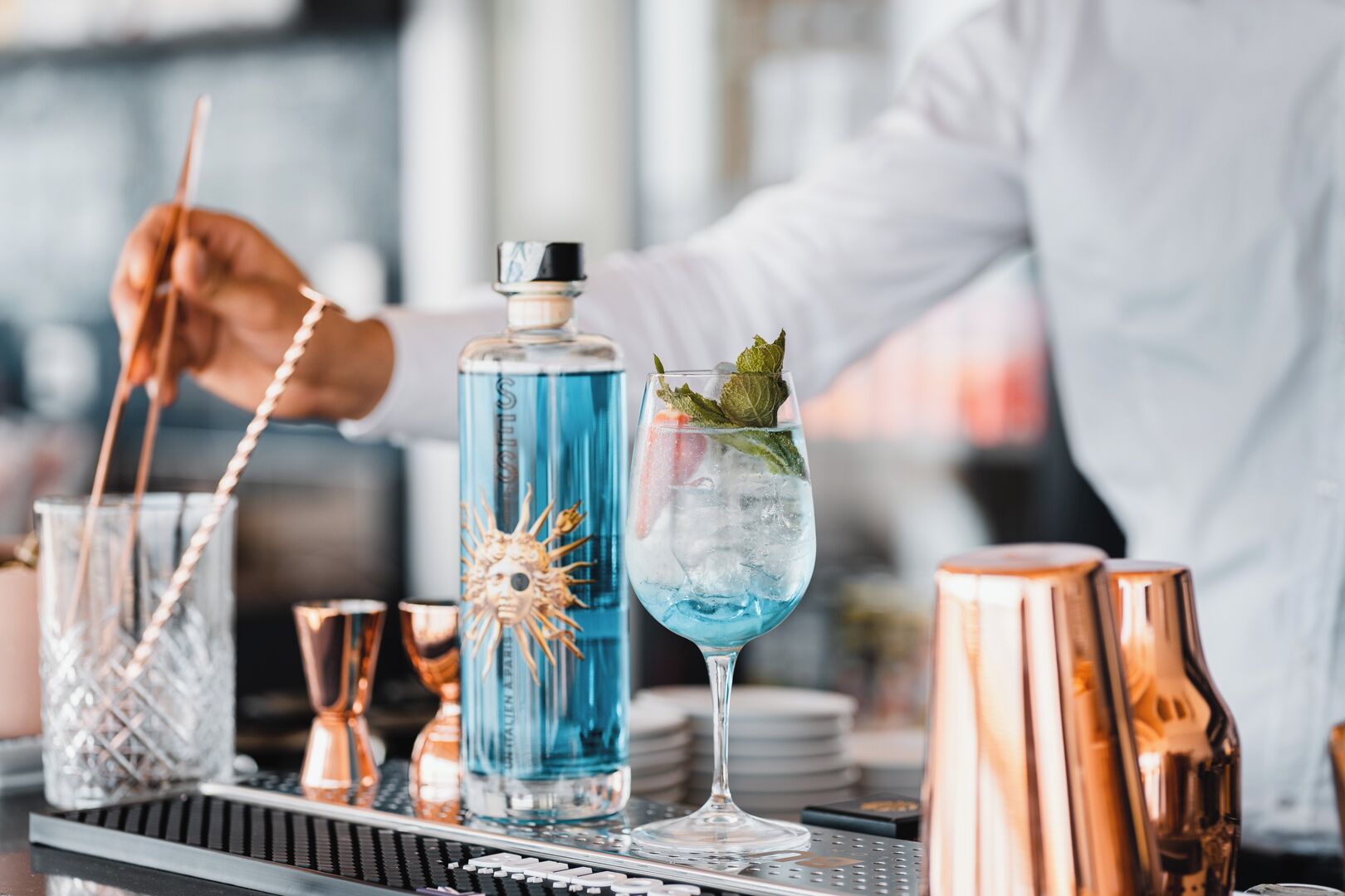 Un bancone bar con un barista che prepara un cocktail, una bottiglia di liquido blu e shaker per cocktail in rame.